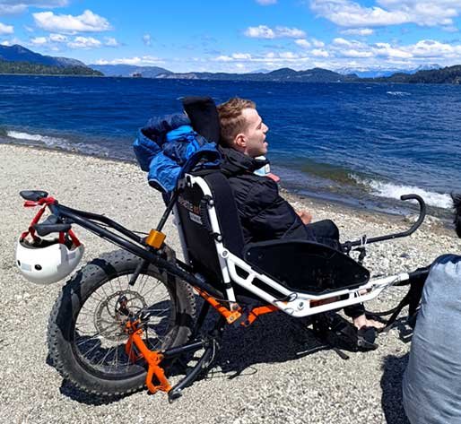 Persona usuaria de silla de ruedas adaptada para playa disfrutando de la costa y el paisaje del lago en Bariloche.